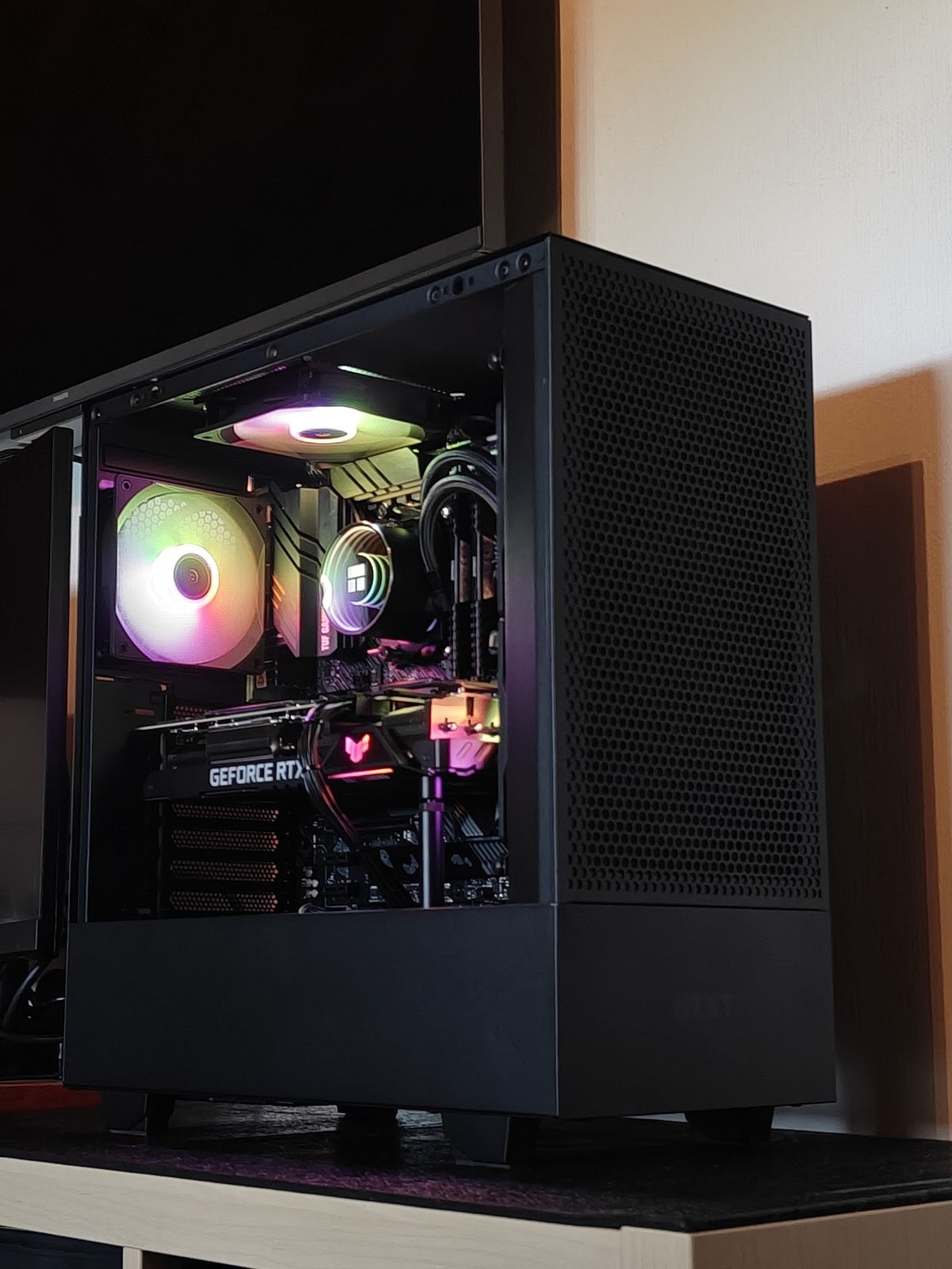 Zwarte meshkast met RGB-fans en een GeForce RTX, onder een televisie
